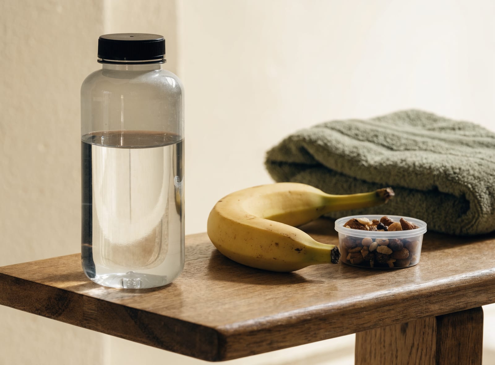 Water bottle, banana, and a container of trail mix on a wooden bench beside a towel